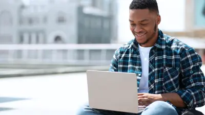 A smiling young man sitting outdoors with a laptop on his lap, symbolizing flexibility and connectivity.