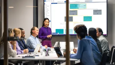 Team sits at a meeting table and woman stands at the front of the screen with a presentation
